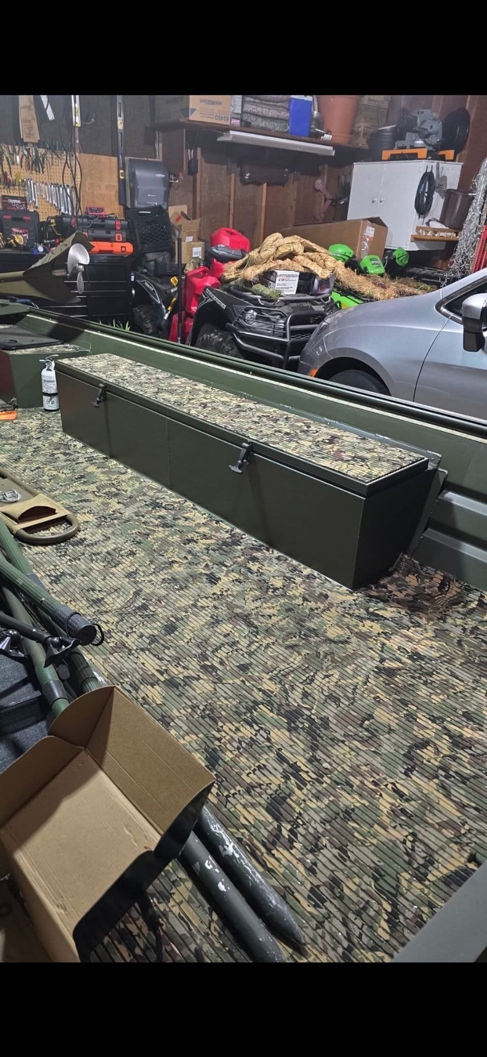 Camouflage storage box on textured floor in a garage filled with tools and vehicles.