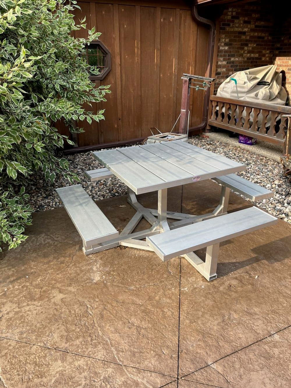 Gray picnic table with benches beside a wooden wall and greenery in a backyard setting.