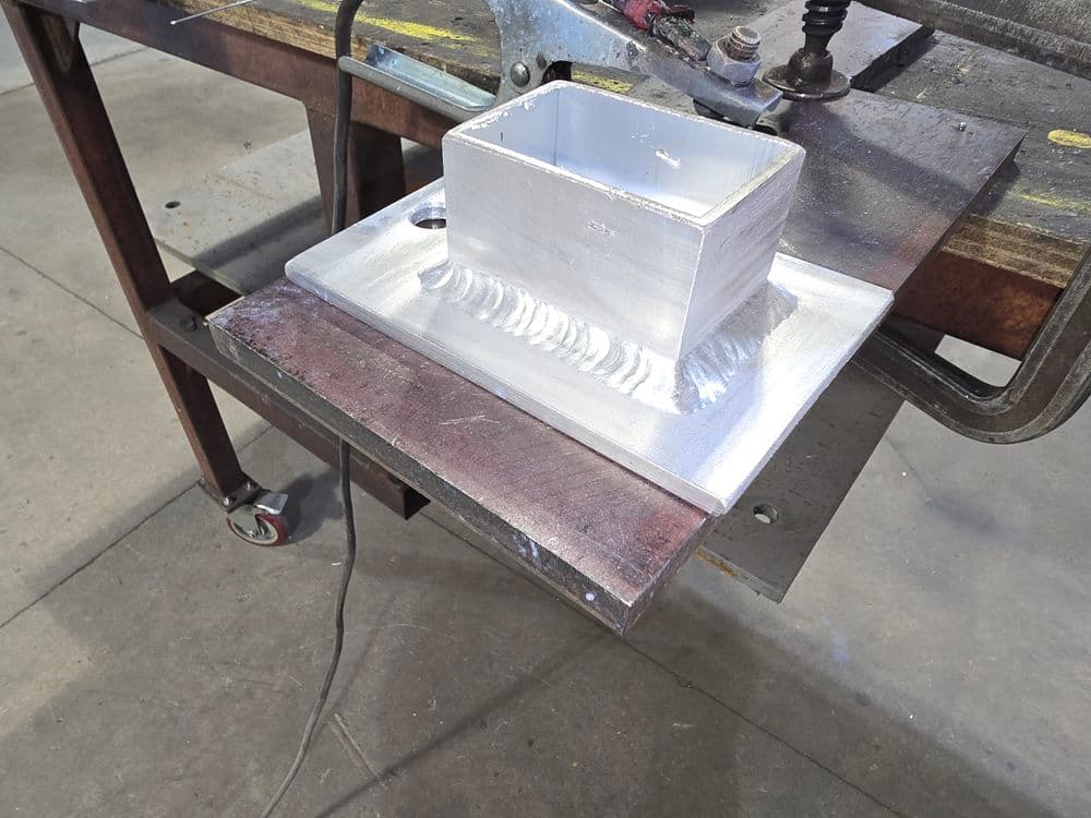 Aluminum block on a metal workbench with welding seams in a workshop setting.
